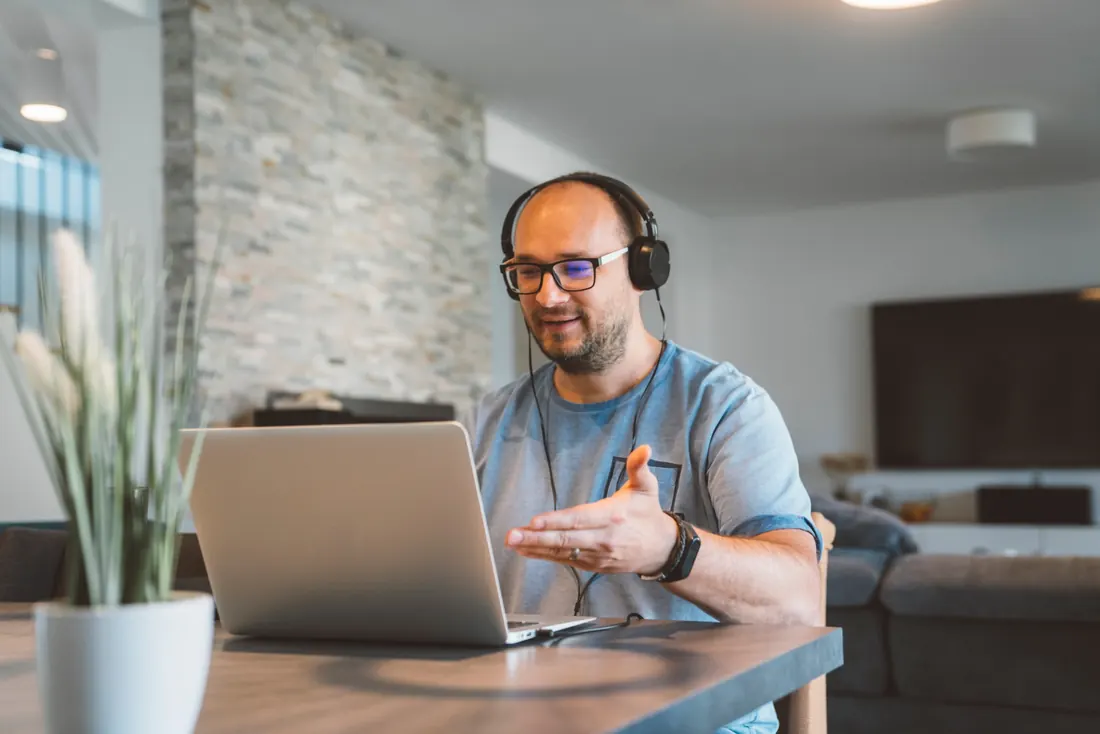 Mann mit Kopfhörern im Videogespräch am Laptop, zu Hause am Esstisch